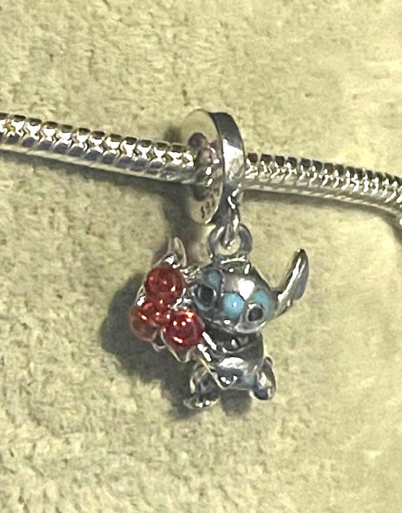 Pode incluir: Um berloque de prata com o personagem Stitch de Lilo & Stitch, segurando um cacho de bagas vermelhas. O berloque tem olhos azuis e est&aacute; preso a uma pulseira de prata.