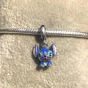 Puede incluir: Un charm de esmalte azul y rosa con el personaje Stitch de la pel&iacute;cula de Disney Lilo & Stitch. El charm est&aacute; suspendido de una pulsera de metal plateado. El charm tiene orejas grandes y una cara detallada.
