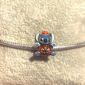 May include: A blue and orange charm featuring a cartoon character wearing a hat and sitting on a pumpkin. The charm is attached to a silver chain bracelet. The charm is a Halloween-themed accessory.