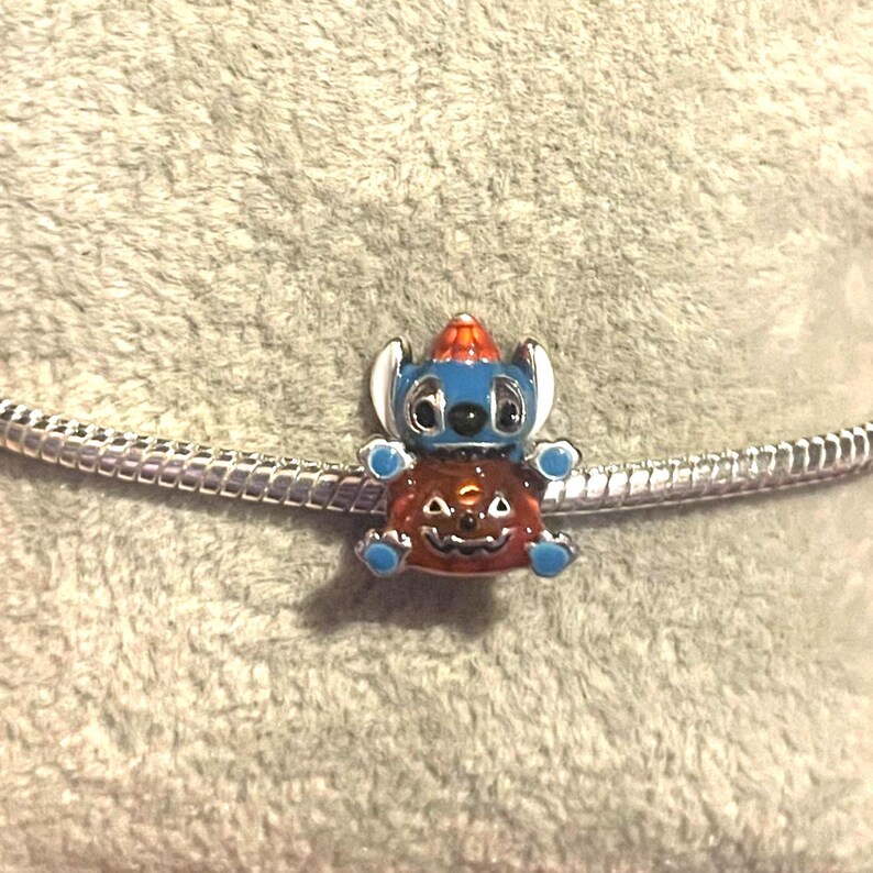 May include: A blue and orange charm featuring a cartoon character wearing a hat and sitting on a pumpkin. The charm is attached to a silver chain bracelet. The charm is a Halloween-themed accessory.