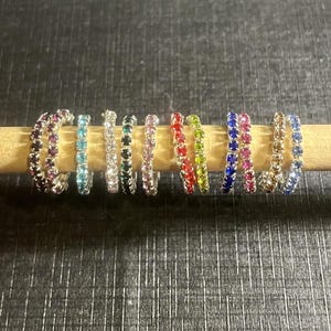 May include: A collection of fourteen small hoop earrings arranged in a row, each featuring a single line of sparkling gemstones in various colors including red, blue, green, pink, clear, and yellow, set in a metallic silver-toned frame.