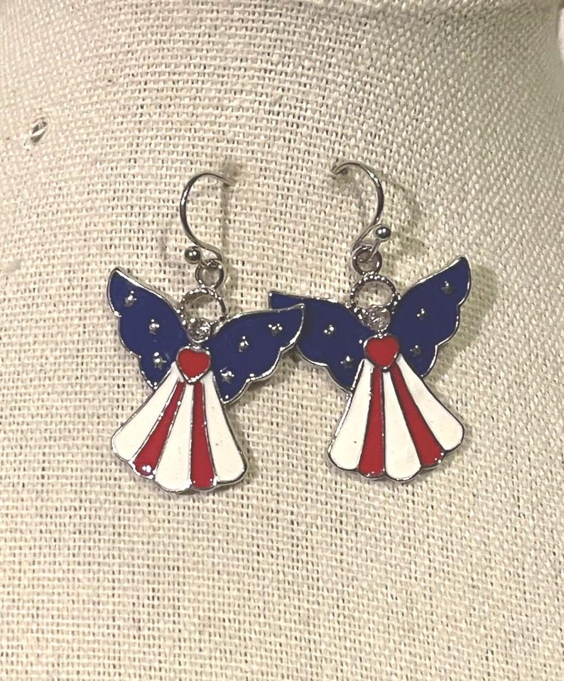 K&ouml;nnte beinhalten: Ein Paar patriotische Engels-Ohrringe. Jeder Ohrring hat blaue Fl&uuml;gel mit wei&szlig;en Sternen, ein rotes Herz und ein wei&szlig;-rot gestreiftes Kleid. Die Ohrringe haben silberfarbene Haken.