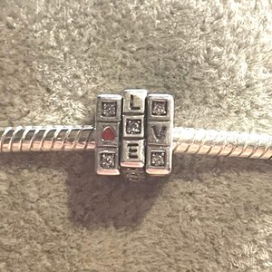 May include: A silver charm with the word "LOVE" spelled out in individual blocks. The "L", "O", and "V" blocks are square with a diamond pattern. The "O" has a red heart. The charm is on a silver bracelet.
