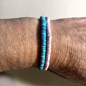 May include: Two beaded bracelets, one light blue and one pink, are stacked on a wrist. The bracelets are made of small, round beads. The light blue bracelet has a marbled effect. The pink bracelet is a solid color.