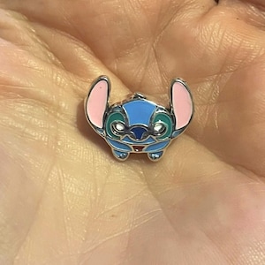 Może przedstawiać: Mały, niebieski, r&oacute;żowy i zielony emaliowany charms w kształcie postaci z kresk&oacute;wki. Charms ma srebrną metalową obw&oacute;dkę. Postać ma duże uszy, okrągłą głowę i małe czerwone usta. Charms jest szczeg&oacute;łowy.