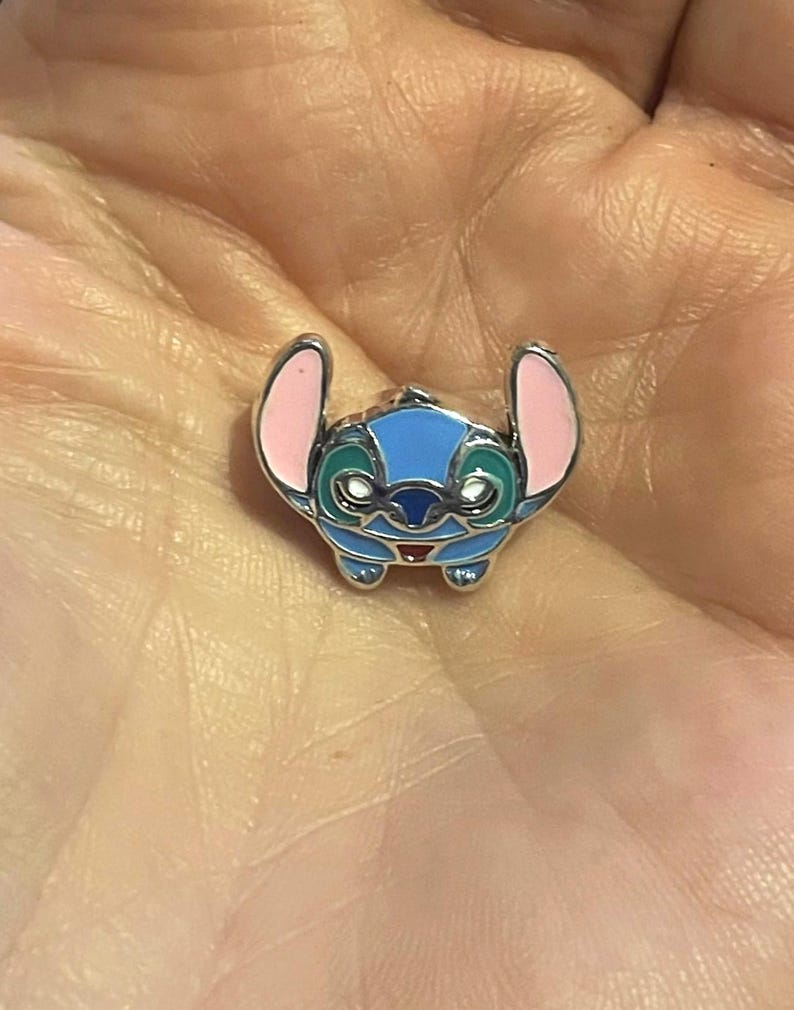 Może przedstawiać: Mały, niebieski, r&oacute;żowy i zielony emaliowany charms w kształcie postaci z kresk&oacute;wki. Charms ma srebrną metalową obw&oacute;dkę. Postać ma duże uszy, okrągłą głowę i małe czerwone usta. Charms jest szczeg&oacute;łowy.