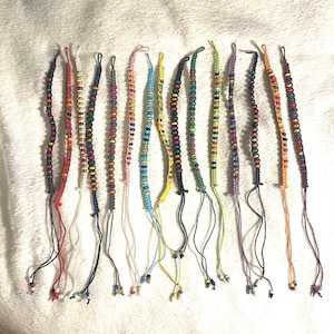 May include: A collection of colorful handmade friendship bracelets. Each bracelet features a unique combination of beads in various colors, including red, orange, yellow, green, blue, and purple. The bracelets are made with knotted string and have dangling ends.