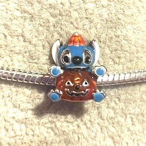 May include: A charm featuring a blue character with white ears and a red and orange hat, wearing a jack-o'-lantern costume. The charm is on a silver bracelet. The charm has blue accents and black eyes.