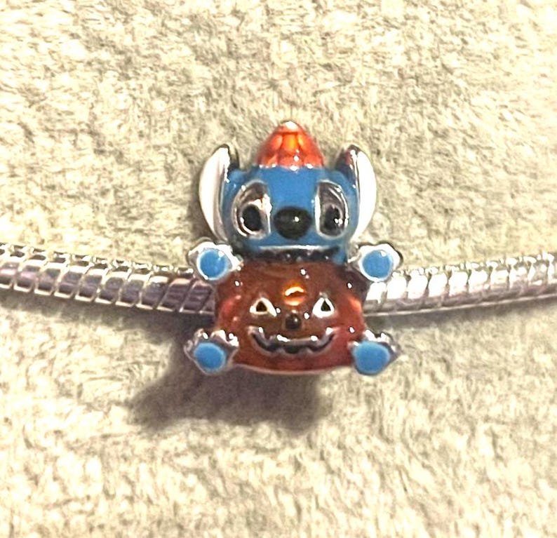 May include: A charm featuring a blue character with white ears and a red and orange hat, wearing a jack-o'-lantern costume. The charm is on a silver bracelet. The charm has blue accents and black eyes.