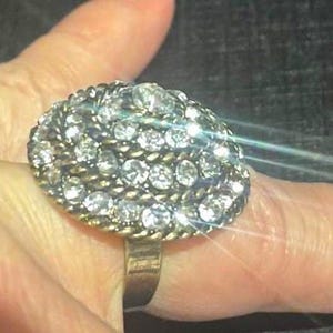 May include: A large, oval-shaped ring with a cluster of clear, sparkling rhinestones. The ring has a gold-toned band and a textured, rope-like border around the rhinestone cluster. The ring is a statement piece.