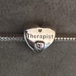 May include: A silver heart-shaped charm with the word "Therapist" engraved on it. The charm has a small heart-shaped detail at the top and a heart-shaped gem at the bottom. The charm is on a silver chain.