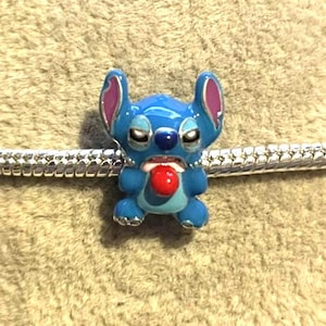 Peut inclure: Un charm bleu &eacute;maill&eacute; repr&eacute;sentant le personnage Stitch de Lilo & Stitch. Le charm a une langue rouge, des oreilles roses et est fix&eacute; &agrave; un bracelet argent&eacute;. Le charm est d&eacute;taill&eacute; et color&eacute;.
