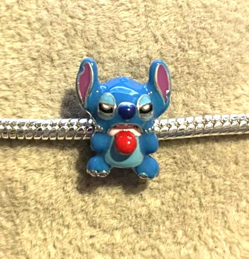 Peut inclure: Un charm bleu &eacute;maill&eacute; repr&eacute;sentant le personnage Stitch de Lilo & Stitch. Le charm a une langue rouge, des oreilles roses et est fix&eacute; &agrave; un bracelet argent&eacute;. Le charm est d&eacute;taill&eacute; et color&eacute;.