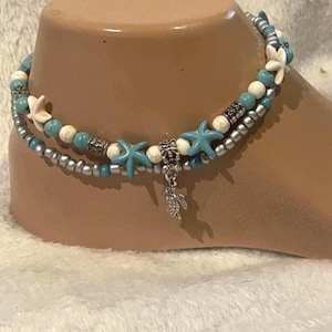 May include: Two-strand anklet with turquoise, white, and silver beads, accented with blue starfish and a silver turtle charm. The anklet is displayed on a tan foot form, suggesting beach or summer wear.