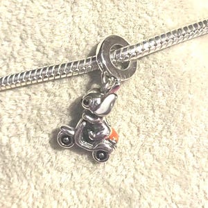 Puede incluir: Un charm de plata con un conejito montado en un scooter. El conejito tiene la cara blanca, orejas rosas y lleva una chaqueta negra. El scooter tiene un asiento naranja y ruedas plateadas. El charm est&aacute; unido a una pulsera de plata.