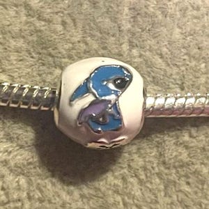 Pode incluir: Uma conta de berloque com um design de personagem de desenho animado azul e roxo em um fundo branco. O berloque &eacute; preso a uma pulseira de prata. A conta tem um acabamento brilhante e provavelmente &eacute; feita de metal.