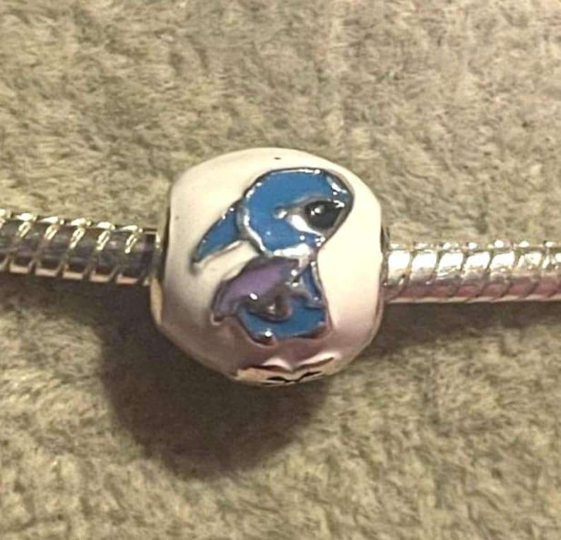 Pode incluir: Uma conta de berloque com um design de personagem de desenho animado azul e roxo em um fundo branco. O berloque &eacute; preso a uma pulseira de prata. A conta tem um acabamento brilhante e provavelmente &eacute; feita de metal.
