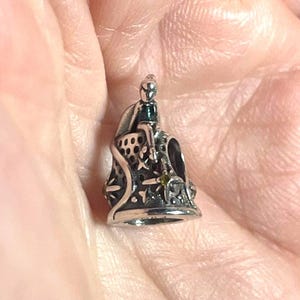 May include: A silver-toned charm with a seated figure wearing a green-colored garment. The charm has intricate cut-out designs and small green accents. The charm is held in a hand.