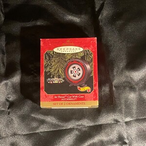 May include: Red and gold box with a Christmas ornament inside. The ornament is a black tire with a silver rim and a Hot Wheels logo. The box says "Keepsake Ornament" and "Jet Threat™ Car With Case HOT WHEELS™ SET OF 2 ORNAMENTS".
