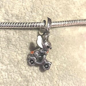 Puede incluir: Un charm de plata con un rat&oacute;n de dibujos animados montado en un scooter. El rat&oacute;n tiene grandes orejas blancas y un asiento naranja. El charm est&aacute; unido a una pulsera de plata.