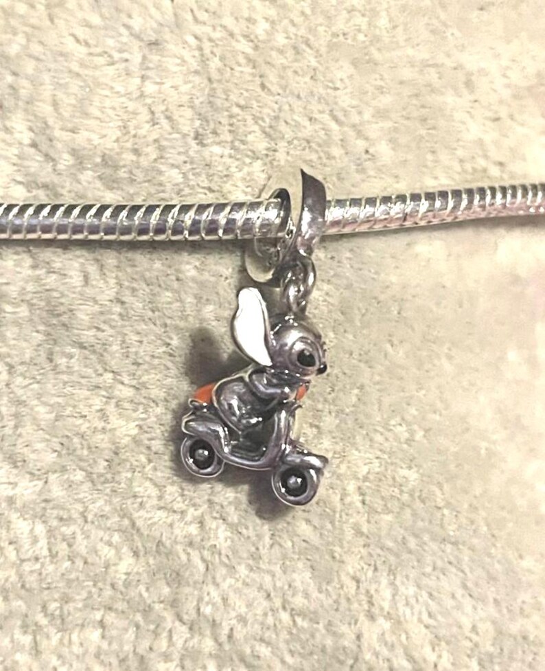 Puede incluir: Un charm de plata con un rat&oacute;n de dibujos animados montado en un scooter. El rat&oacute;n tiene grandes orejas blancas y un asiento naranja. El charm est&aacute; unido a una pulsera de plata.