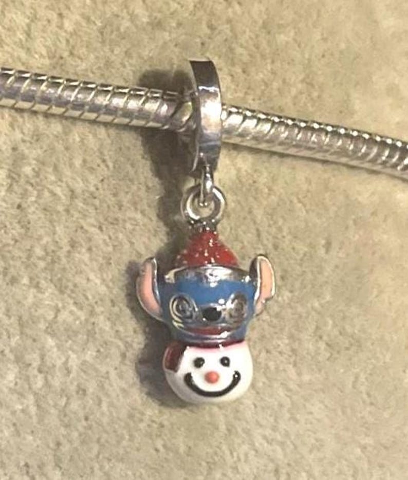 Puede incluir: Un charm de plata con un personaje azul y blanco con un gorro y bufanda rojos, que se asemeja a un mu&ntilde;eco de nieve. El charm est&aacute; unido a una pulsera de plata. El personaje tiene orejas rosas y una cara sonriente.