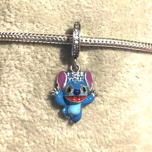 May include: A blue and silver charm featuring a cartoon character with the text "I SEE YOU!". The charm has a blue body, pink ears, and a red mouth. It is attached to a silver chain.