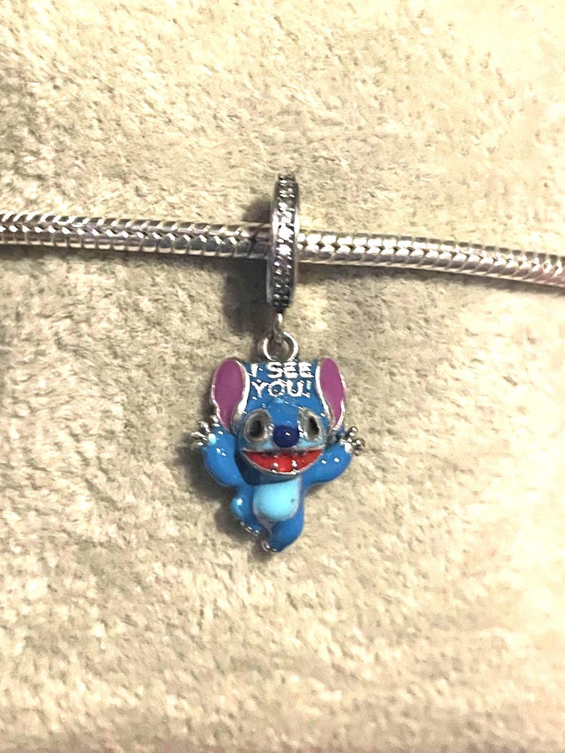 May include: A blue and silver charm featuring a cartoon character with the text "I SEE YOU!". The charm has a blue body, pink ears, and a red mouth. It is attached to a silver chain.