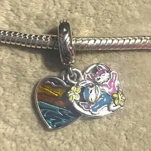 May include: A silver charm with two pendants. One pendant is a heart-shaped design with a sunset scene. The other pendant features a cartoon image of a donkey and a rabbit. The charm is attached to a silver bracelet.