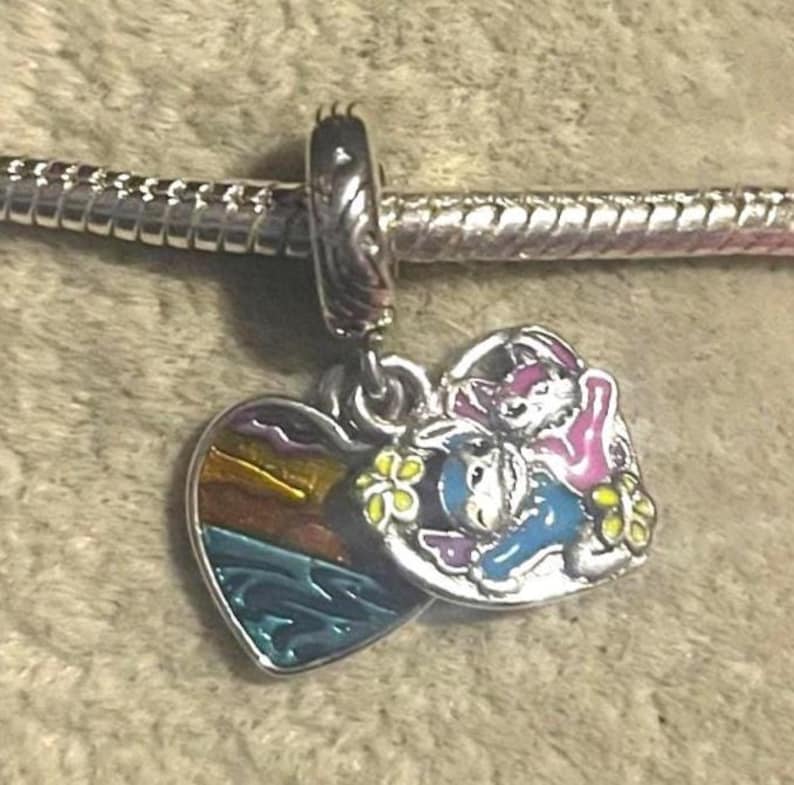 May include: A silver charm with two pendants. One pendant is a heart-shaped design with a sunset scene. The other pendant features a cartoon image of a donkey and a rabbit. The charm is attached to a silver bracelet.