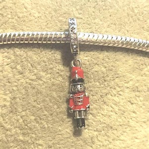 May include: A silver charm featuring a miniature red and black toy soldier. The soldier wears a red uniform and a black hat. The charm is attached to a silver bracelet with a decorative clasp.