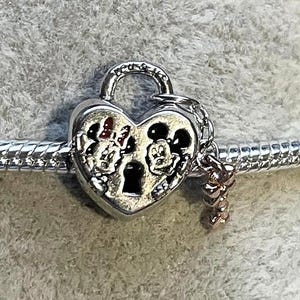 May include: A silver heart-shaped charm with Mickey and Minnie Mouse designs. The charm is a padlock design with a small dangling charm. The charm is on a silver bracelet.