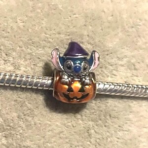 Peut inclure: Un charm repr&eacute;sentant un personnage bleu avec des oreilles roses et un chapeau violet, perch&eacute; sur une citrouille orange. Le charm est fix&eacute; &agrave; un bracelet argent&eacute;, pour Halloween ou les fans de Disney.