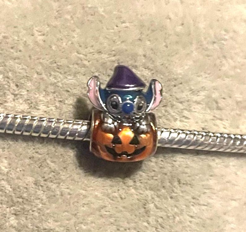 Peut inclure: Un charm repr&eacute;sentant un personnage bleu avec des oreilles roses et un chapeau violet, perch&eacute; sur une citrouille orange. Le charm est fix&eacute; &agrave; un bracelet argent&eacute;, pour Halloween ou les fans de Disney.
