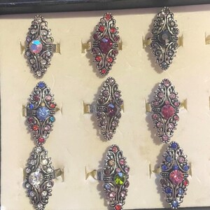 May include: Nine ornate rings with oval-shaped, multi-colored gemstones. The rings have a silver-tone metal base with intricate floral designs. The gemstones are various colors, including red, blue, pink, and clear, set in a decorative pattern.