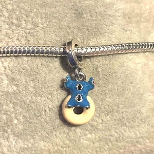 May include: A blue and white enamel charm featuring a cartoon character design. The charm is attached to a silver chain. The charm is in the shape of a blue character on a white donut.