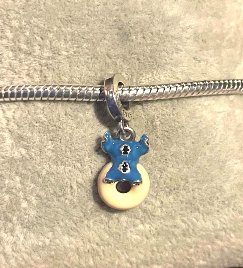 May include: A blue and white enamel charm featuring a cartoon character design. The charm is attached to a silver chain. The charm is in the shape of a blue character on a white donut.