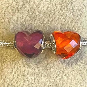 May include: Two heart-shaped charms, one deep purple and the other orange, are displayed on a silver bracelet. The charms have a faceted design, catching the light. The bracelet has a snake chain design.
