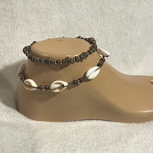 May include: Two anklets on a foot form. One anklet features small brown beads and gold-colored accents. The other anklet has white seashells and brown beads. The anklets are displayed on a beige foot form against a white background.