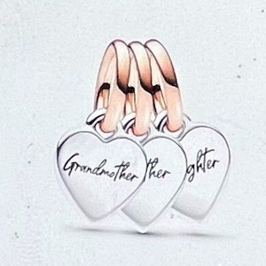 May include: Three silver heart-shaped charms with the words "Grandmother", "Mother", and "Daughter" inscribed on them. The charms are connected by three rose gold rings.
