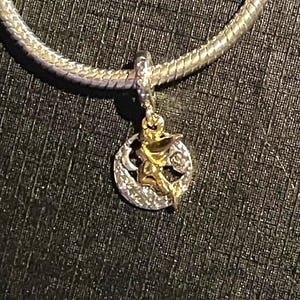 May include: A gold and silver charm pendant featuring a cherub design within a circle, accented with small crystals. The charm is suspended from a silver snake chain necklace. The pendant has a celestial theme.