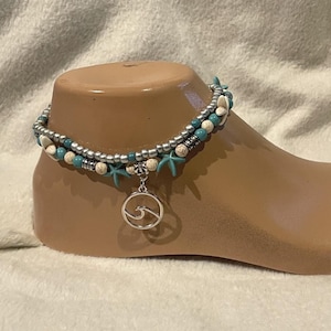 May include: Ankle bracelet featuring turquoise, white, and silver beads, accented with starfish charms. A silver wave pendant hangs from the bracelet. The jewelry is displayed on a foot-shaped display.