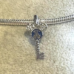 May include: A silver charm in the shape of a key with a Mickey Mouse head design. The key's head features a blue enamel background with a castle silhouette. The Mickey Mouse ears and key details are embellished with clear crystals.