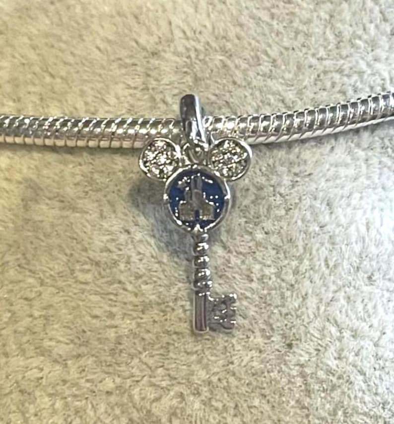 May include: A silver charm in the shape of a key with a Mickey Mouse head design. The key's head features a blue enamel background with a castle silhouette. The Mickey Mouse ears and key details are embellished with clear crystals.