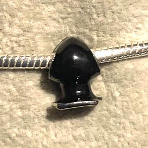 May include: A black, glossy charm bead with a rounded top and a flared base. The bead is attached to a silver-colored, snake chain bracelet. The charm has a smooth, polished finish.