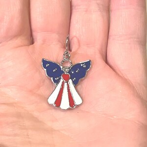 K&ouml;nnte beinhalten: Ein patriotischer Engel-Anh&auml;nger mit einem silberfarbenen Haken. Der Engel hat blaue Fl&uuml;gel mit wei&szlig;en Sternen, ein rotes Herz und ein wei&szlig;-rotes Kleid. Ein kleines, dekoratives Accessoire.