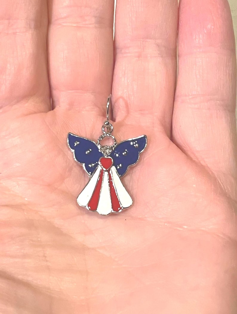 K&ouml;nnte beinhalten: Ein patriotischer Engel-Anh&auml;nger mit einem silberfarbenen Haken. Der Engel hat blaue Fl&uuml;gel mit wei&szlig;en Sternen, ein rotes Herz und ein wei&szlig;-rotes Kleid. Ein kleines, dekoratives Accessoire.