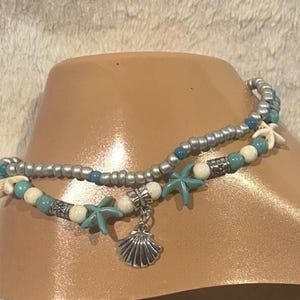May include: Two-strand anklet with silver beads, turquoise starfish, and white beads. A silver seashell charm hangs from the lower strand. The anklet is displayed on a tan foot form, suggesting beach or summer wear.