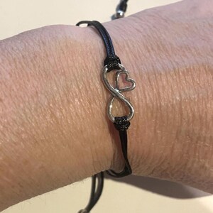 May include: Black cord bracelet featuring a silver-colored infinity symbol with a pink heart accent. The bracelet is adjustable and has a sliding knot closure. A simple and elegant piece of jewelry.