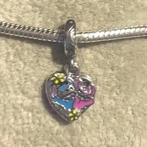 May include: A silver charm with a heart-shaped pendant featuring a blue and pink cartoon character design. The charm is attached to a silver chain. Yellow flowers are scattered around the characters. The charm is likely for a bracelet or necklace.